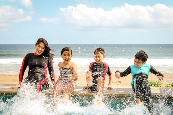  A woman and three children joyfully splashing water while sitting on the edge of a pool, with a beach and ocean in the background.