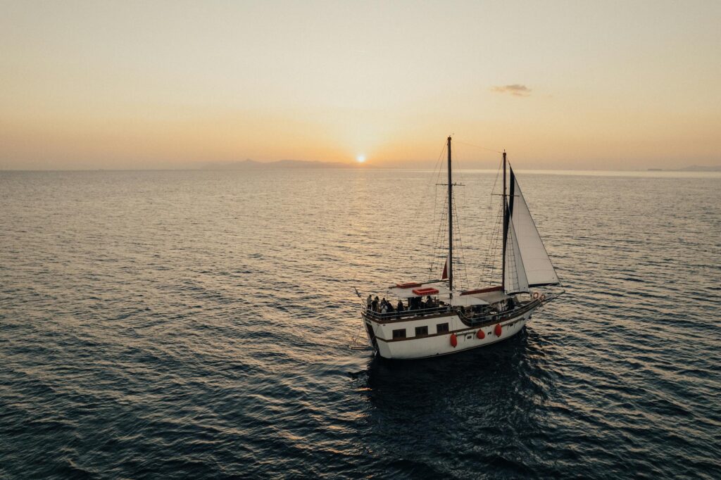 A sailing boat with a white sail on calm waters during sunset, with the sun setting on the horizon and a clear sky