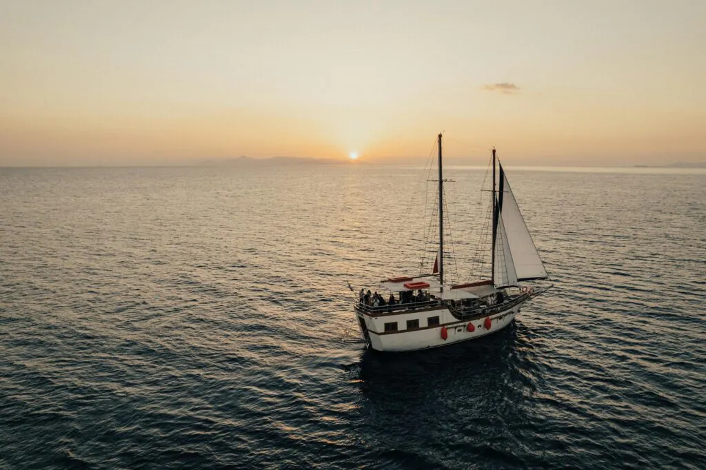 A sailing boat with a white sail on calm waters during sunset, with the sun setting on the horizon and a clear sky