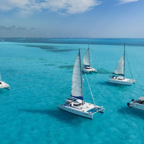 Five white catamarans with sails raised, sailing in clear turquoise waters under a blue sky with scattered clouds.
